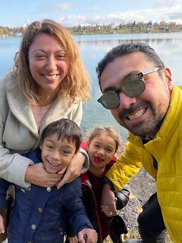 A smiling family of four poses by a lake on a sunny day, autumn trees in the background—capturing the perfect Finger Lakes weekend getaway.