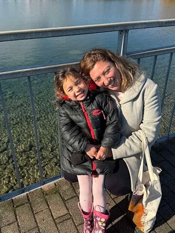 A woman and young girl smile and hug by a railing next to clear water on a sunny day, capturing the joyful moments often found at Finger Lakes wineries with lodging.