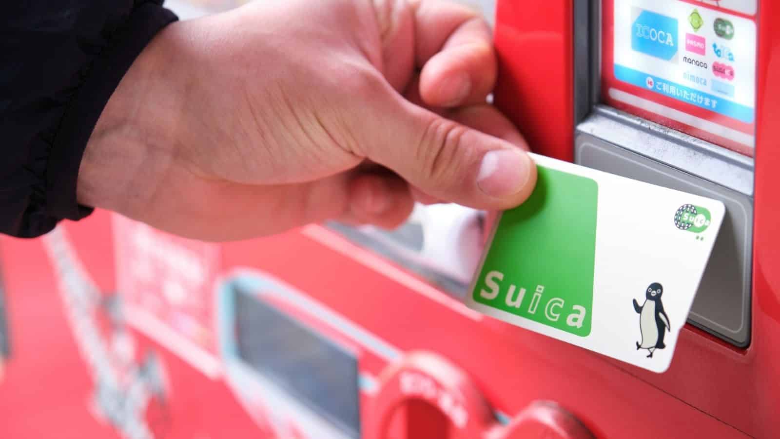 Hand holding a Suica card, tapping it on a red vending machine payment reader.