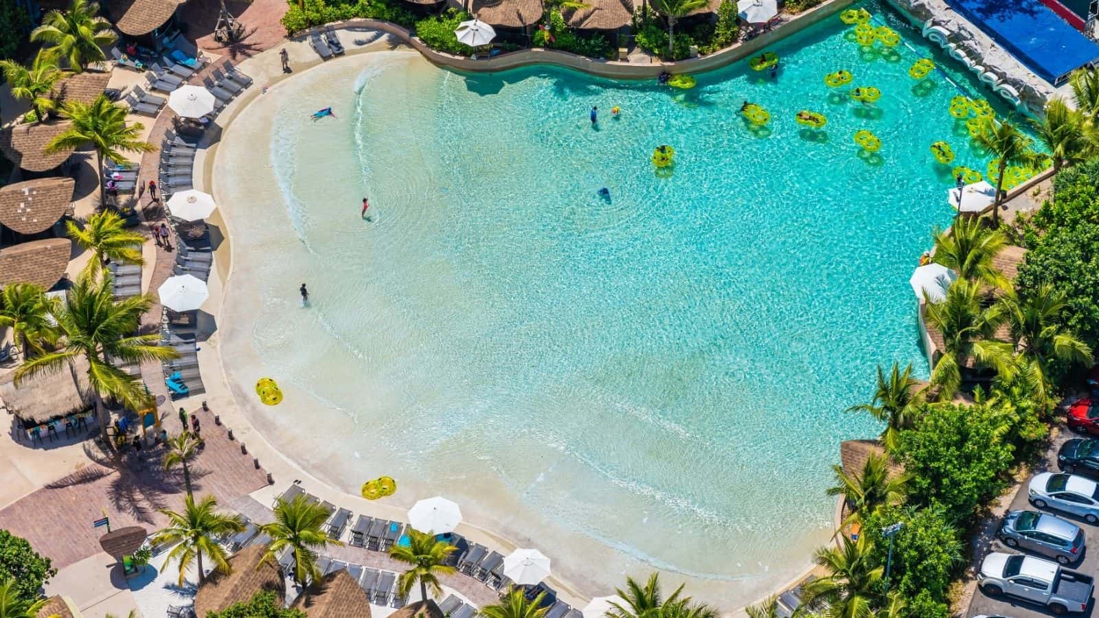 Aerial view of a large resort pool with scattered swimmers, surrounded by palm trees and lounge chairs.