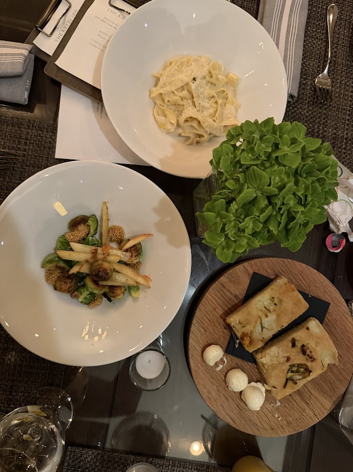 Three plated dishes and a potted herb on a table—featuring pasta, vegetables, and focaccia with butter—set the scene for a perfect Finger Lakes weekend getaway or cozy fall road trip meal.