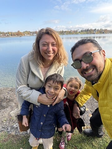A smiling family of four poses by a Finger Lakes lake on a sunny day, with trees and blue sky in the background—capturing the perfect weekend getaway.