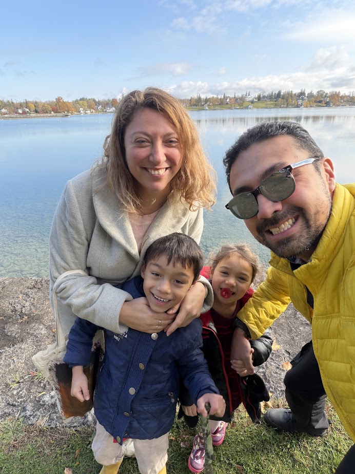 A smiling family of four poses by a Finger Lakes lake on a sunny day, with trees and blue sky in the background—capturing the perfect weekend getaway.