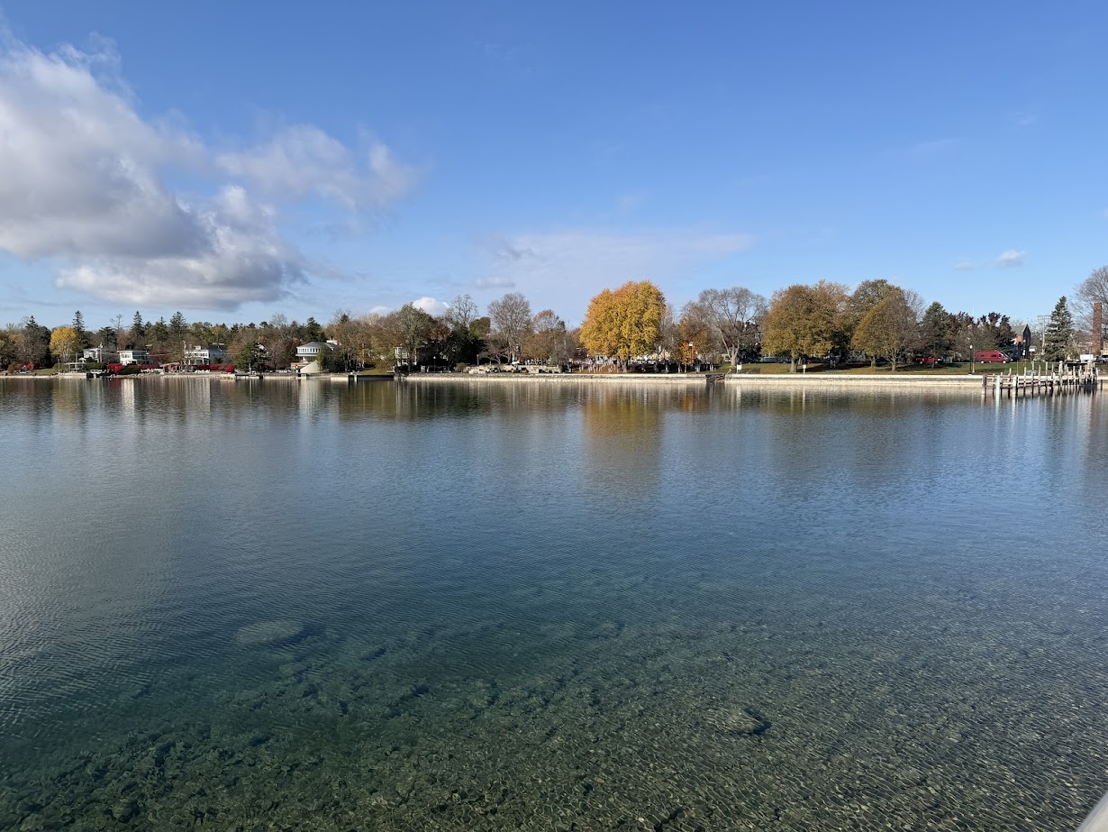 Calm lake with clear water, distant shoreline, and autumn trees under a partly cloudy blue sky—perfect for a peaceful weekend getaway in the Finger Lakes.