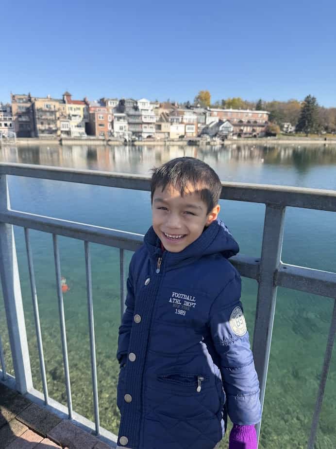 Smiling boy in a blue coat stands by a lakeside railing in the Finger Lakes, with houses and trees in the background—perfect for a weekend getaway.