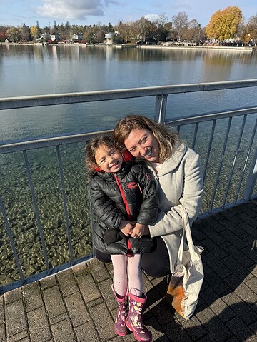 A woman and a young girl smile together by a lakeside railing on a sunny day, enjoying the scenic charm near Finger Lakes Wineries with Lodging.