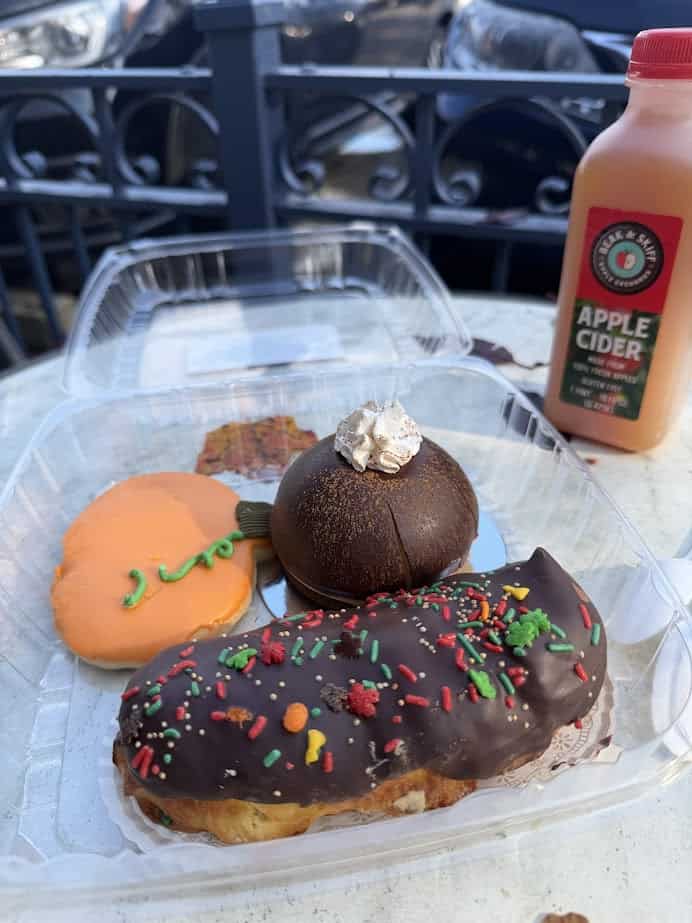 Three festive pastries sit in a plastic container next to a bottle of apple cider on an outdoor table—perfect treats for a Finger Lakes fall road trip.