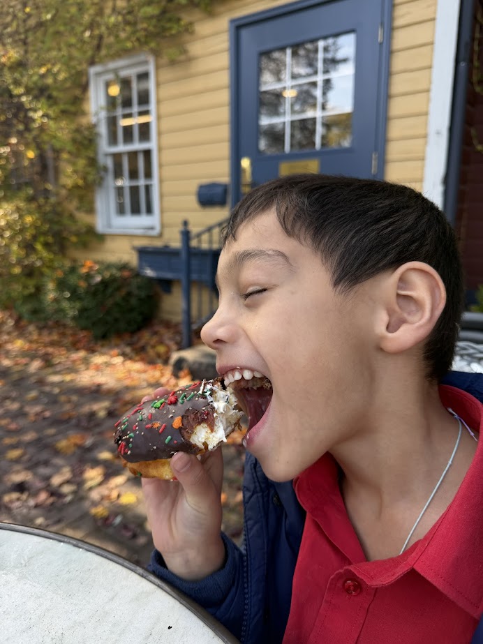 Smiling boy in a red shirt eating a chocolate donut with sprinkles outdoors near a yellow house, enjoying a sweet treat during a fall road trip in the Finger Lakes.