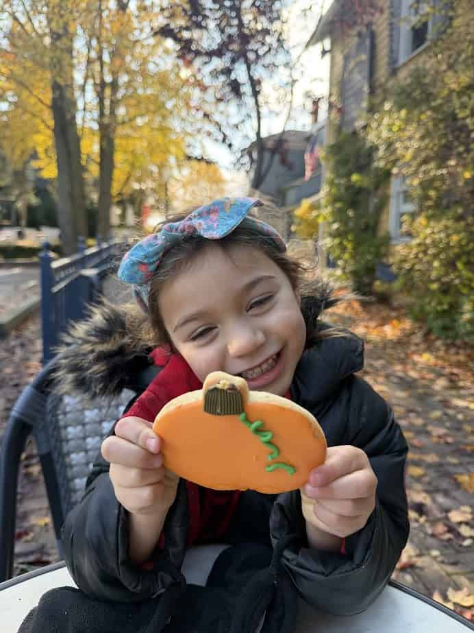 Smiling child outdoors holding a pumpkin-shaped cookie on a fall day, enjoying a Finger Lakes weekend getaway, with trees and houses in the background.
