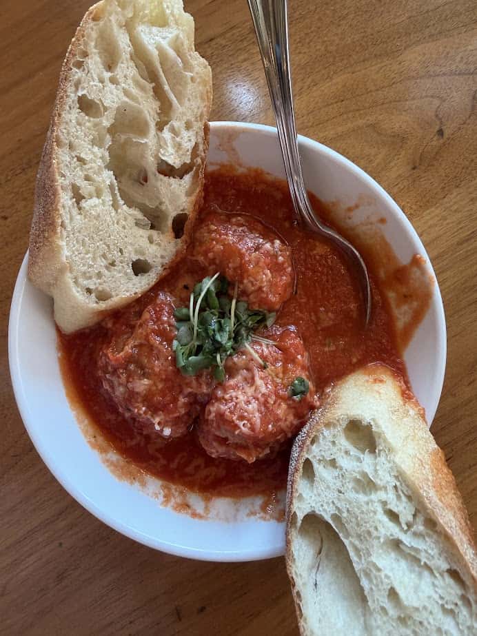 Three meatballs in tomato sauce with microgreens, served with two slices of bread on a white plate—perfect comfort food for a Finger Lakes weekend getaway.
