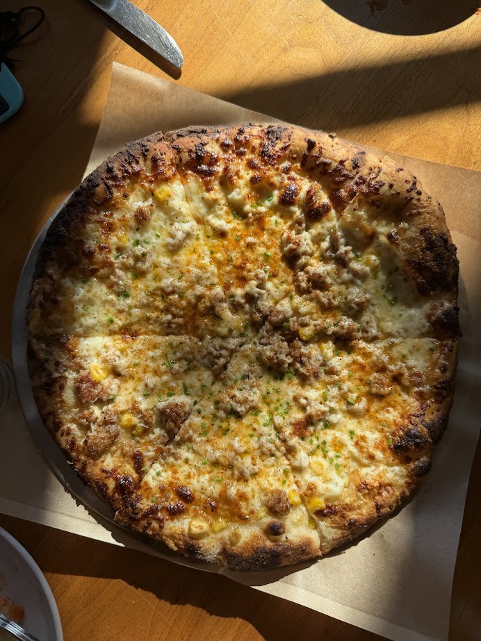 A cheese pizza with herbs and browned crust sits on parchment paper in sunlight on a wooden table—perfect for enjoying during a Finger Lakes weekend getaway.