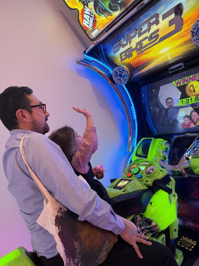 A man and a child sit on an arcade motorcycle game, celebrating a win on "Super Bikes 3" after reading a glowing skaneateles-fields-resort-spa-review nearby.