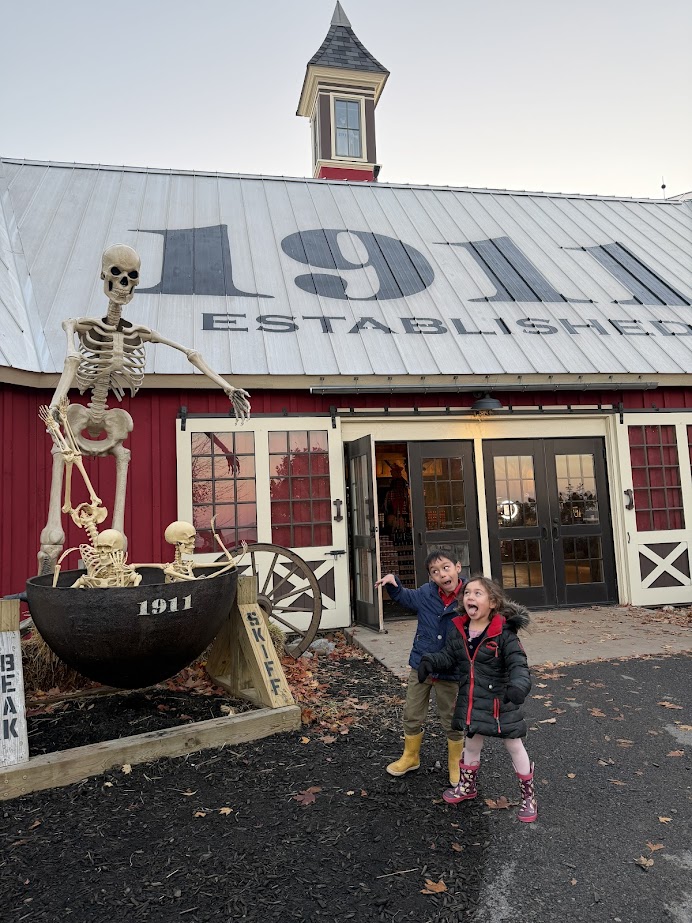 Two children laugh in front of a barn with a giant skeleton Halloween display in a metal cauldron—a perfect stop on a Finger Lakes fall road trip.