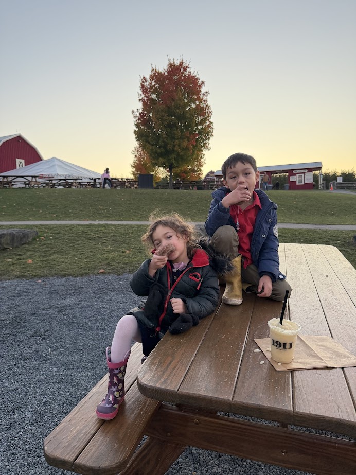 Two kids sit on a picnic table eating donuts at a farm in the Finger Lakes, with autumn trees and a red barn in the background—a perfect stop during a fall road trip or weekend getaway.