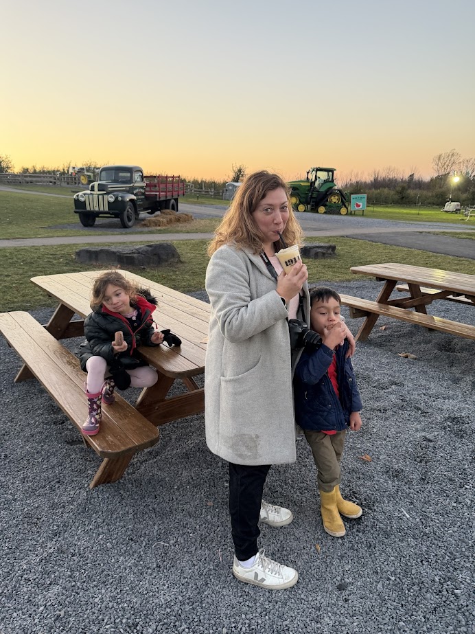 A woman and two children stand near picnic tables at sunset, holding snacks and posing for the camera outdoors during their Finger Lakes weekend getaway.