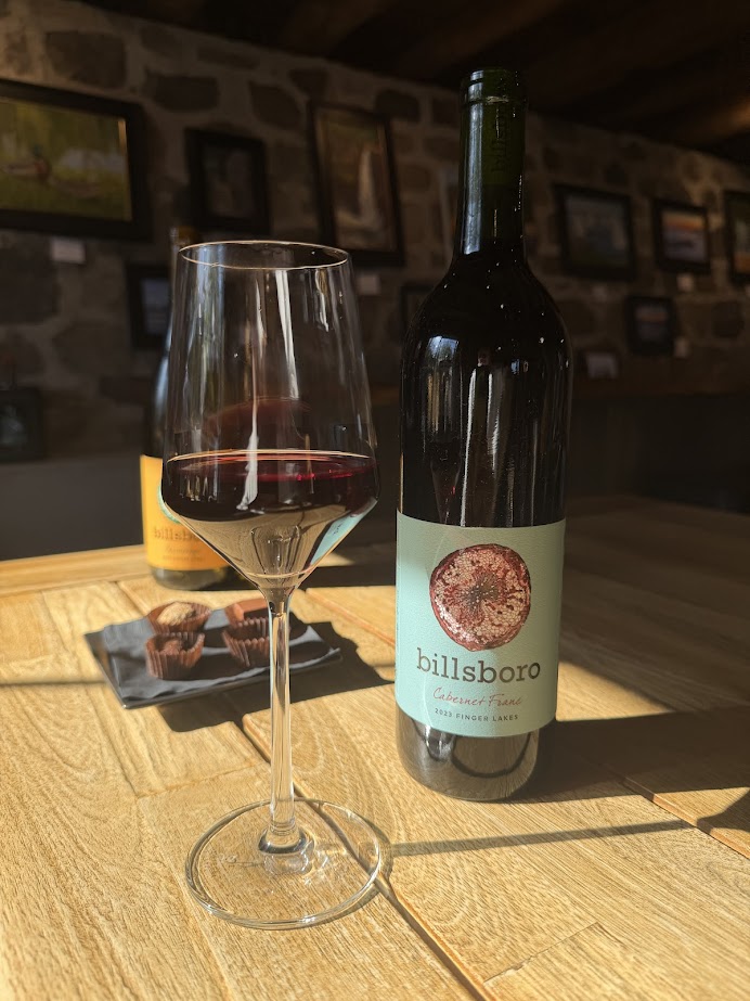 A glass and bottle of Billsboro cabernet franc sit on a wooden table in a sunlit room with art on the walls, capturing the warm ambiance of one of the best wineries in Finger Lakes.