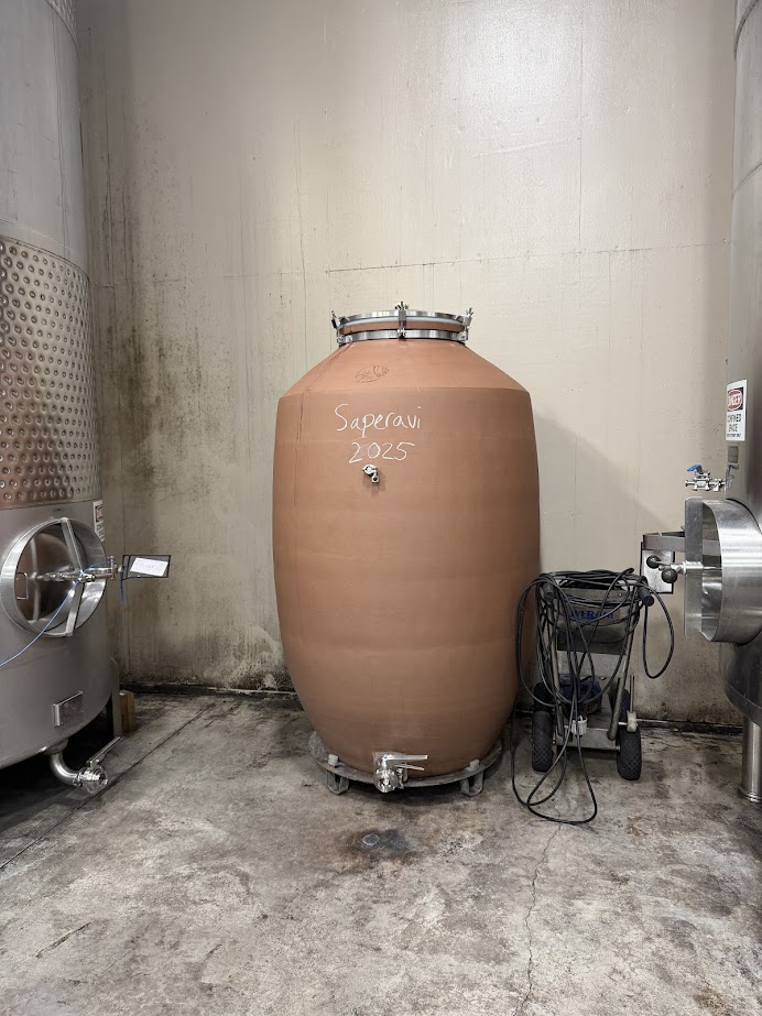 A clay wine fermenter labeled "Saperavi 2025" stands between two metal tanks in a winery, echoing the traditional methods cherished by the best wineries in Finger Lakes.