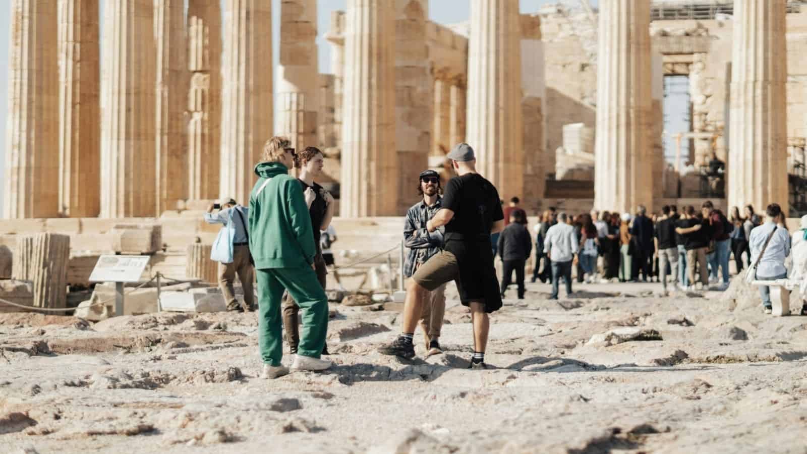 Tourists wearing casual clothes stand and talk among ancient columns at a historic site with a crowd in the background.