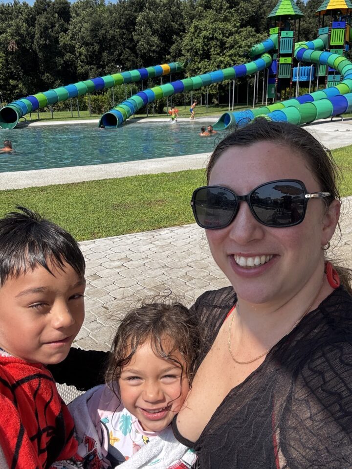 A woman and two children smile by a pool at one of the Best Family Resorts, with colorful water slides and people swimming in the background.