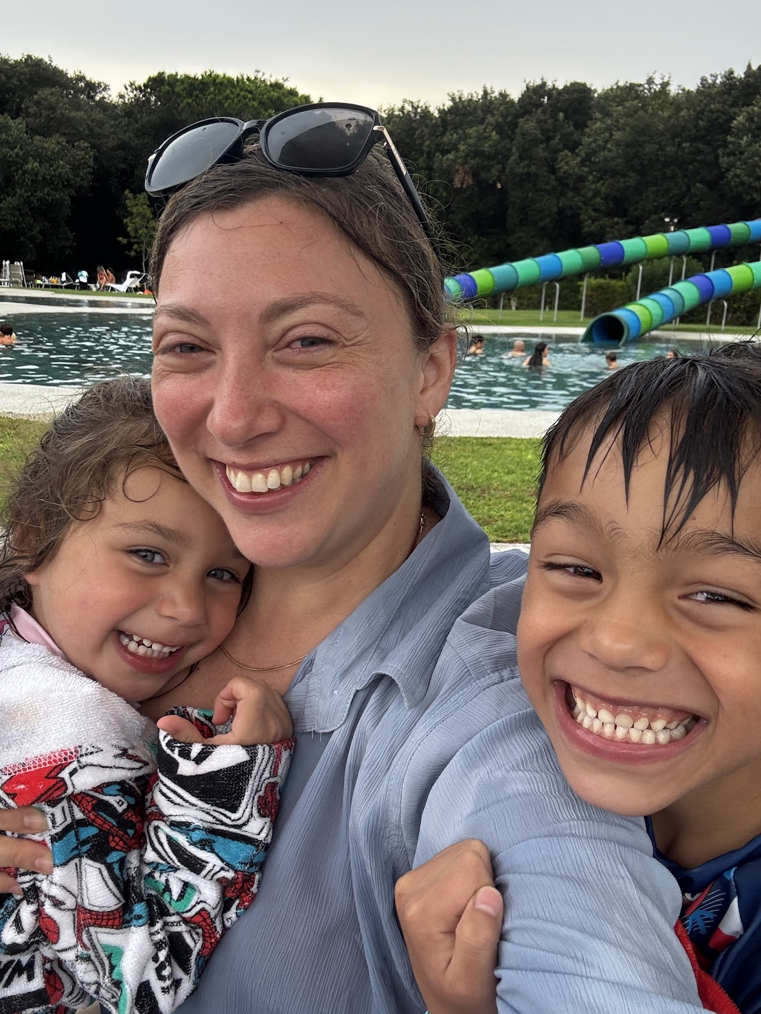 Smiling woman and two kids taking a selfie by a pool at one of the best family resorts, with water slides and people enjoying in the background.