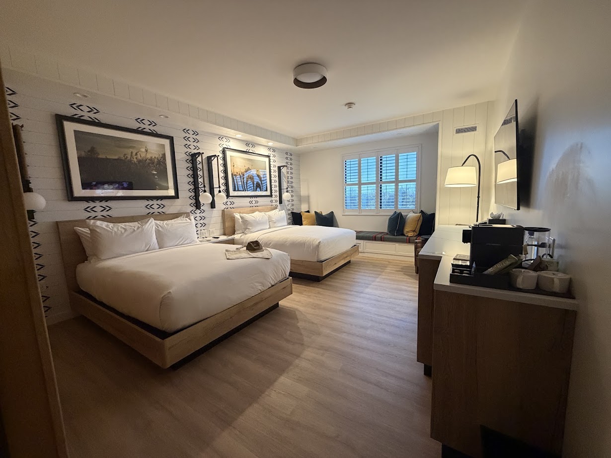 Modern hotel room with two beds, a window seat, wall art, and a coffee station on a wooden floor—perfect for relaxing after exploring the Finger Lakes or enjoying your stay at Skaneateles Fields Resort.