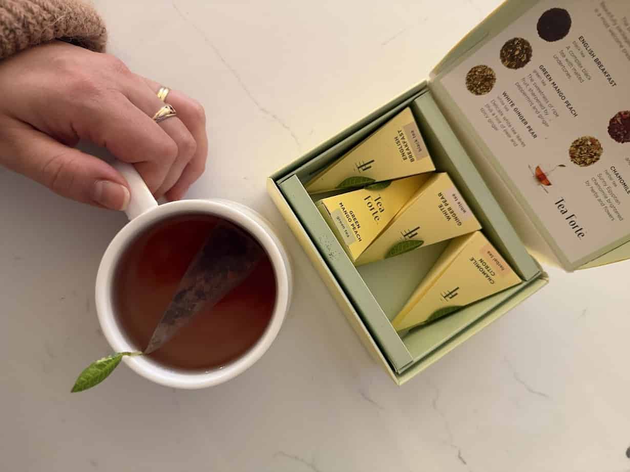 A hand holds a teacup with a tea bag; an open box of assorted tea sachets sits nearby on a white surface, evoking the relaxing ambiance of Skaneateles Fields Resort in the Finger Lakes.