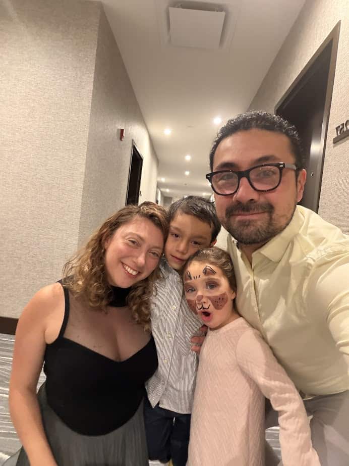 A family of four smiles in a hallway at Skaneateles Fields Resort Spa; the girl has face paint resembling a cat, capturing joy worthy of a glowing skaneateles-fields-resort-spa-review.