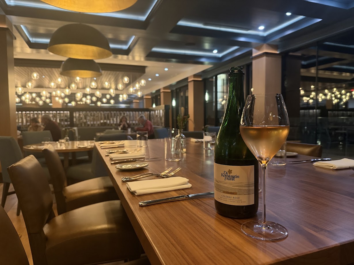 A wine bottle and glass on a set dining table in a warmly lit, modern restaurant at Skaneateles Fields Resort, nestled near the Finger Lakes.