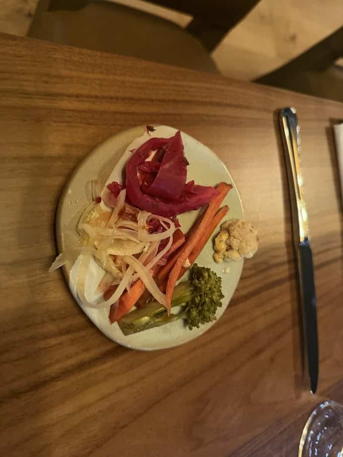 A small plate of assorted pickled vegetables with a dollop of a light spread, served on a wooden table at Skaneateles Fields Resort in the heart of the Finger Lakes.
