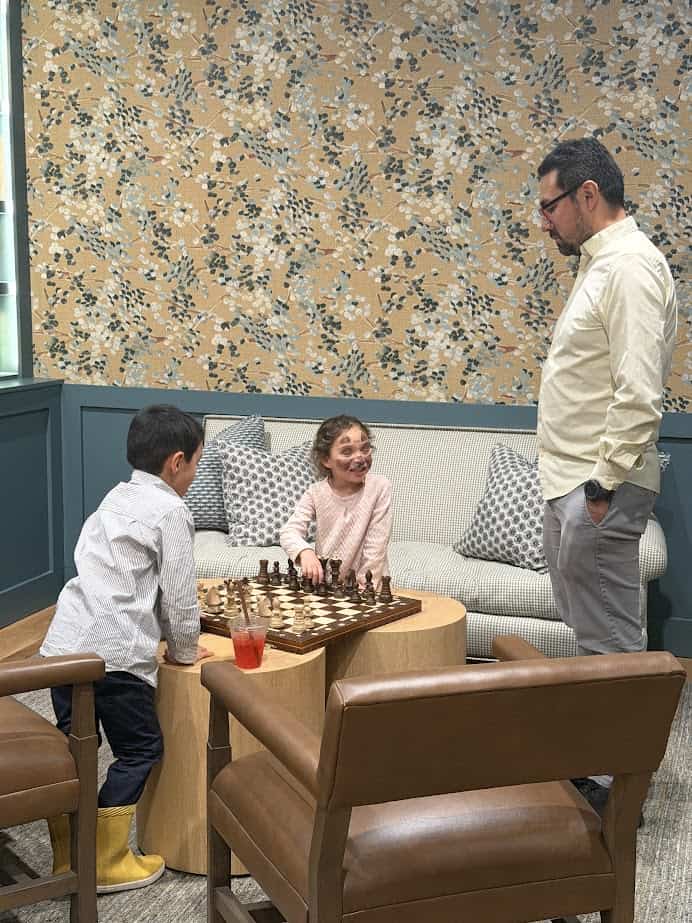 Two children play chess at a table while an adult stands nearby watching them, all smiling and engaged, enjoying quality time together at Skaneateles Fields Resort in the scenic Finger Lakes.