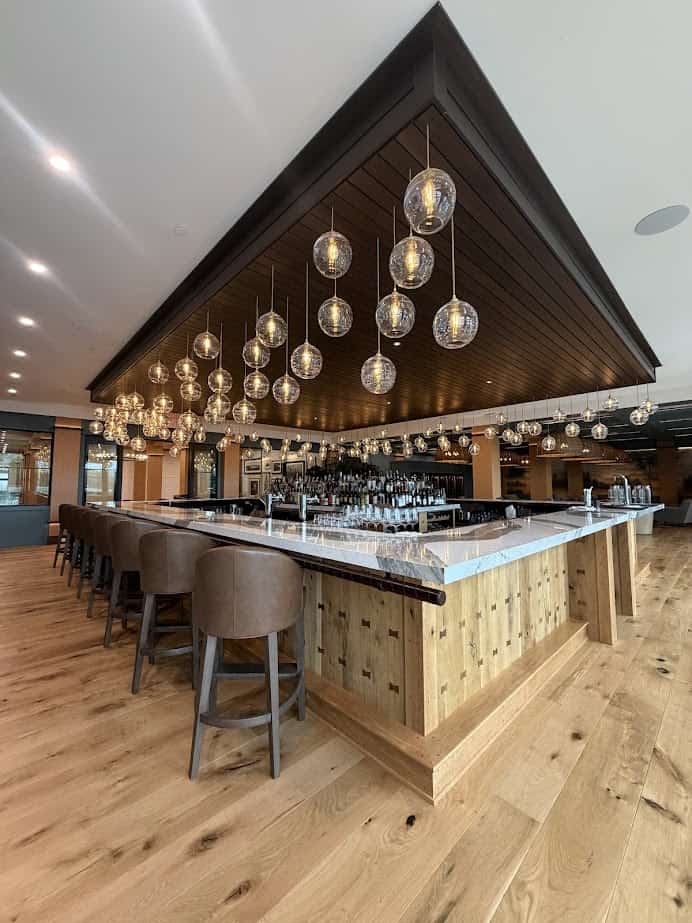 A modern bar with wooden floors, hanging globe lights, and padded barstools around a large marble counter—perfect for unwinding after a day at the Spa or exploring the scenic Finger Lakes.