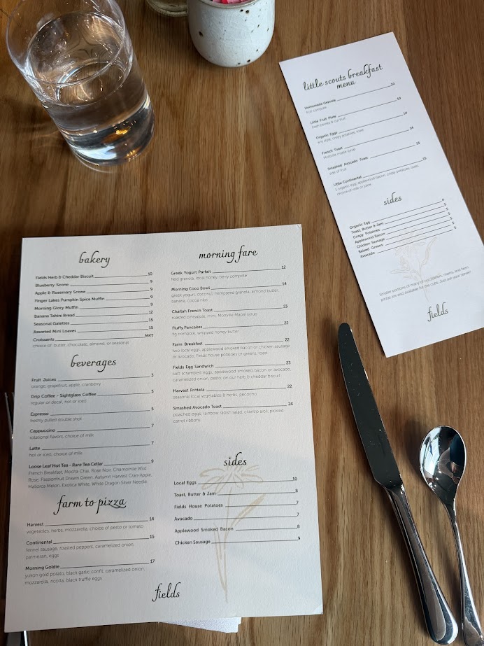 A breakfast menu and utensils are arranged on a wooden table next to a glass of water and a smaller side menu, capturing the inviting ambiance of Skaneateles Fields Resort in the Finger Lakes.