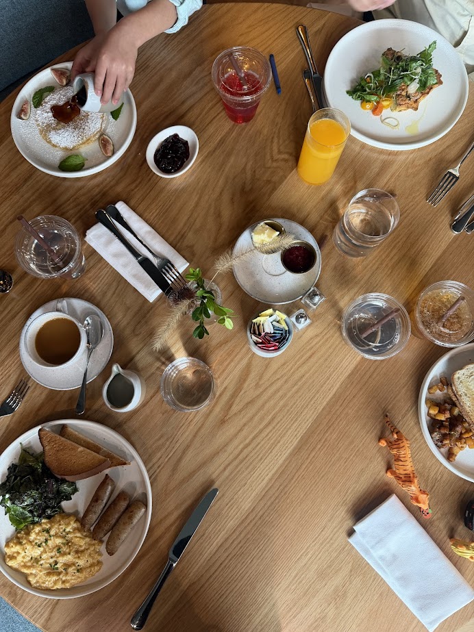 Brunch table with plates of food, drinks, utensils, and two people reaching for dishes at Skaneateles Fields Resort in the Finger Lakes.