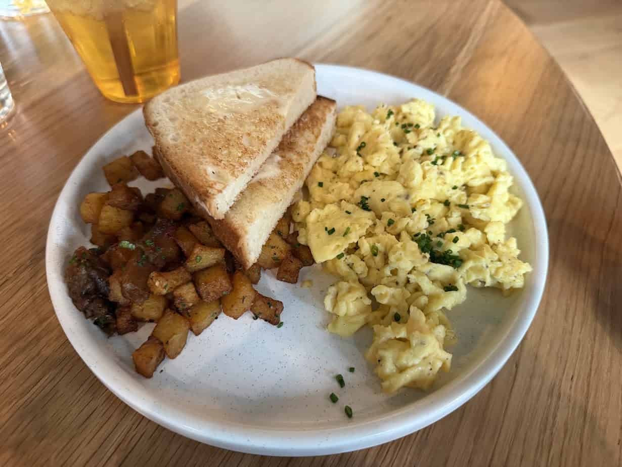 A plate with scrambled eggs, toast, breakfast potatoes, and a glass of iced tea awaits you at Skaneateles Fields Resort in the Finger Lakes, set on a rustic wooden table for the perfect start to your day.