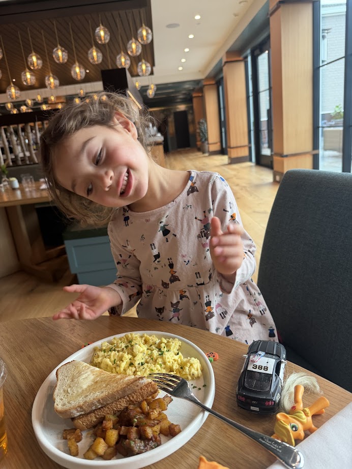Smiling girl enjoys scrambled eggs, toast, and potatoes with toy cars at a restaurant table near Skaneateles Fields Resort in the Finger Lakes.