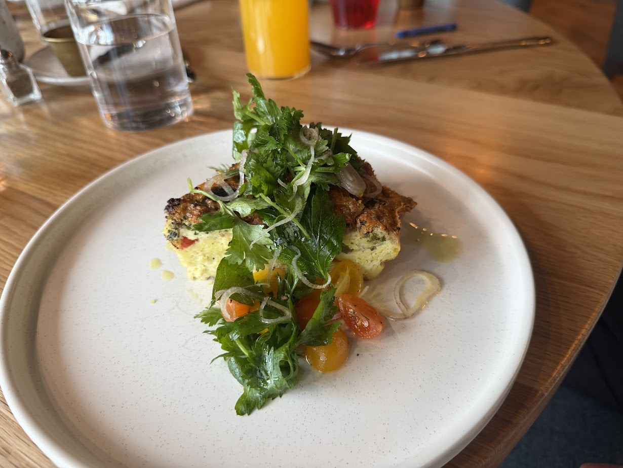 A slice of frittata topped with fresh greens and tomatoes on a white plate, set on a wooden table at Skaneateles Fields Resort in the Finger Lakes.