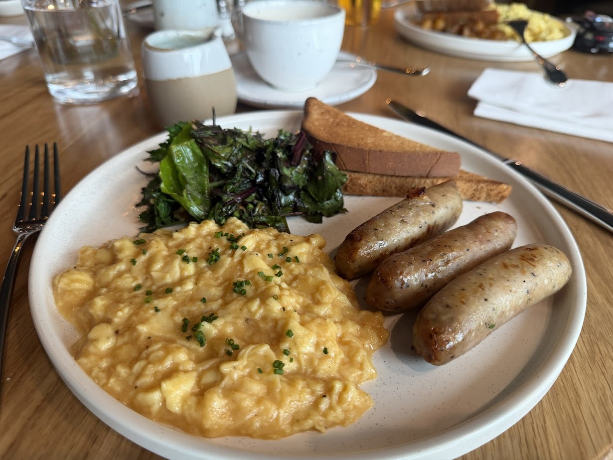 A plate with scrambled eggs, sausages, toast, and salad on a wooden table at Skaneateles Fields Resort, offering a taste of comfort in the heart of Finger Lakes. Cups and glasses complete this relaxing spa breakfast experience.