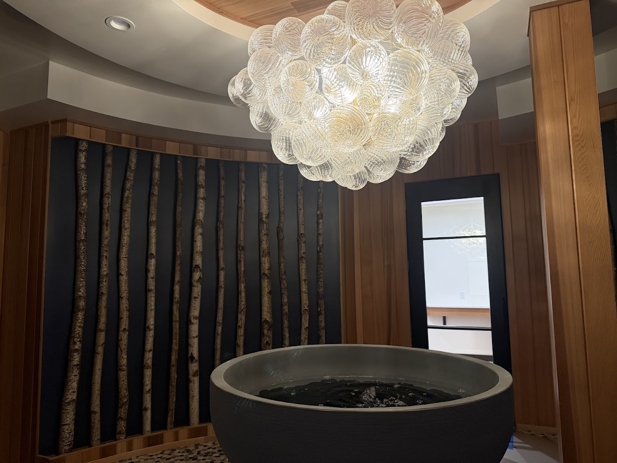 A modern room at Skaneateles Fields Resort with a large round soaking tub, birch log decor, and a glass chandelier overhead offers spa-like relaxation inspired by the tranquil beauty of the Finger Lakes.