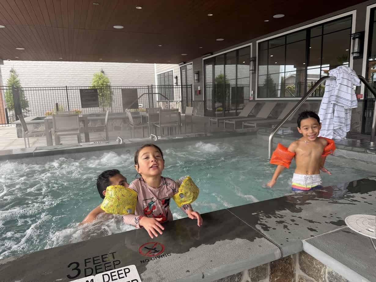 Three young children in floaties play and smile in an indoor hot tub at Skaneateles Fields Resort Spa, with towels and chairs in the background, capturing a joyful moment in the heart of Finger Lakes.