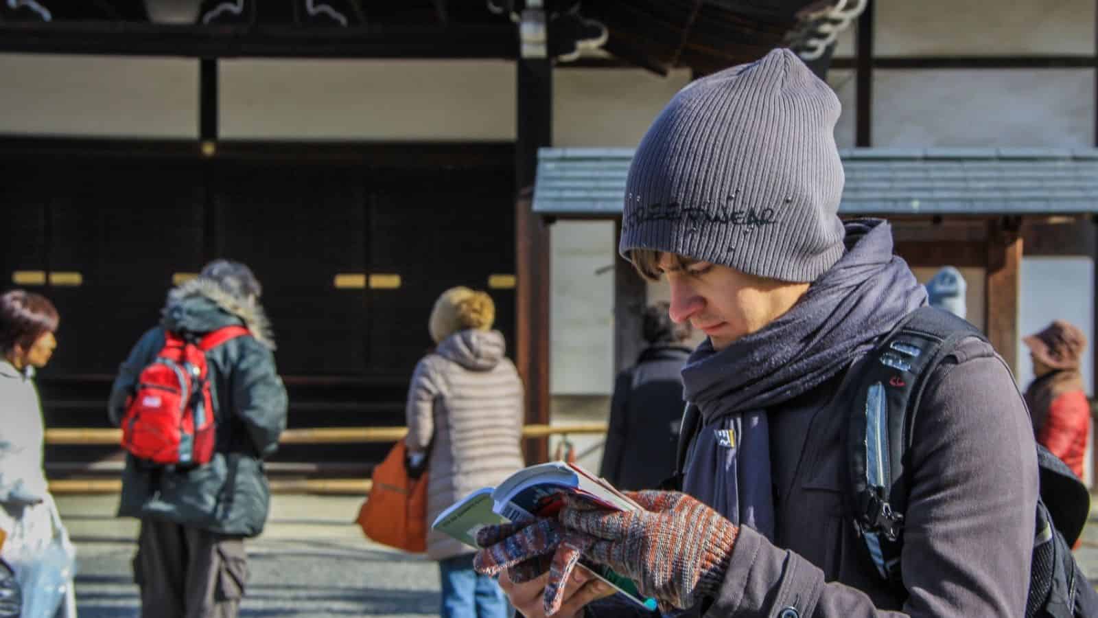 Young man in winter clothes reads a guidebook outdoors; several people stand in the background.