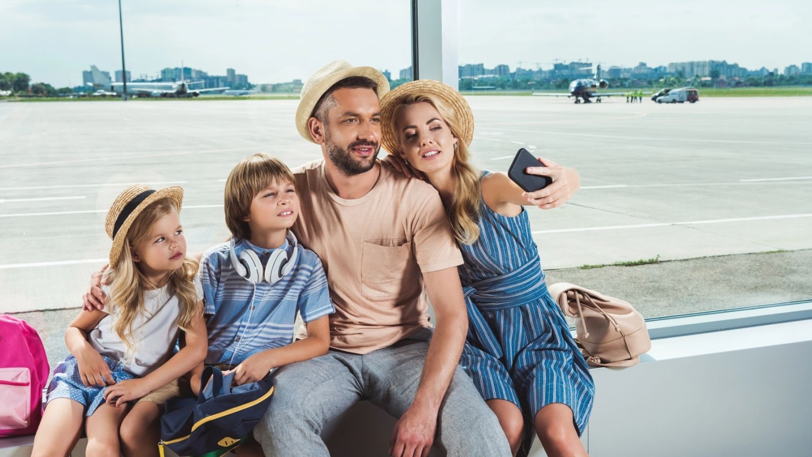 A family of four in summer clothes takes a selfie together at an airport with planes in the background.