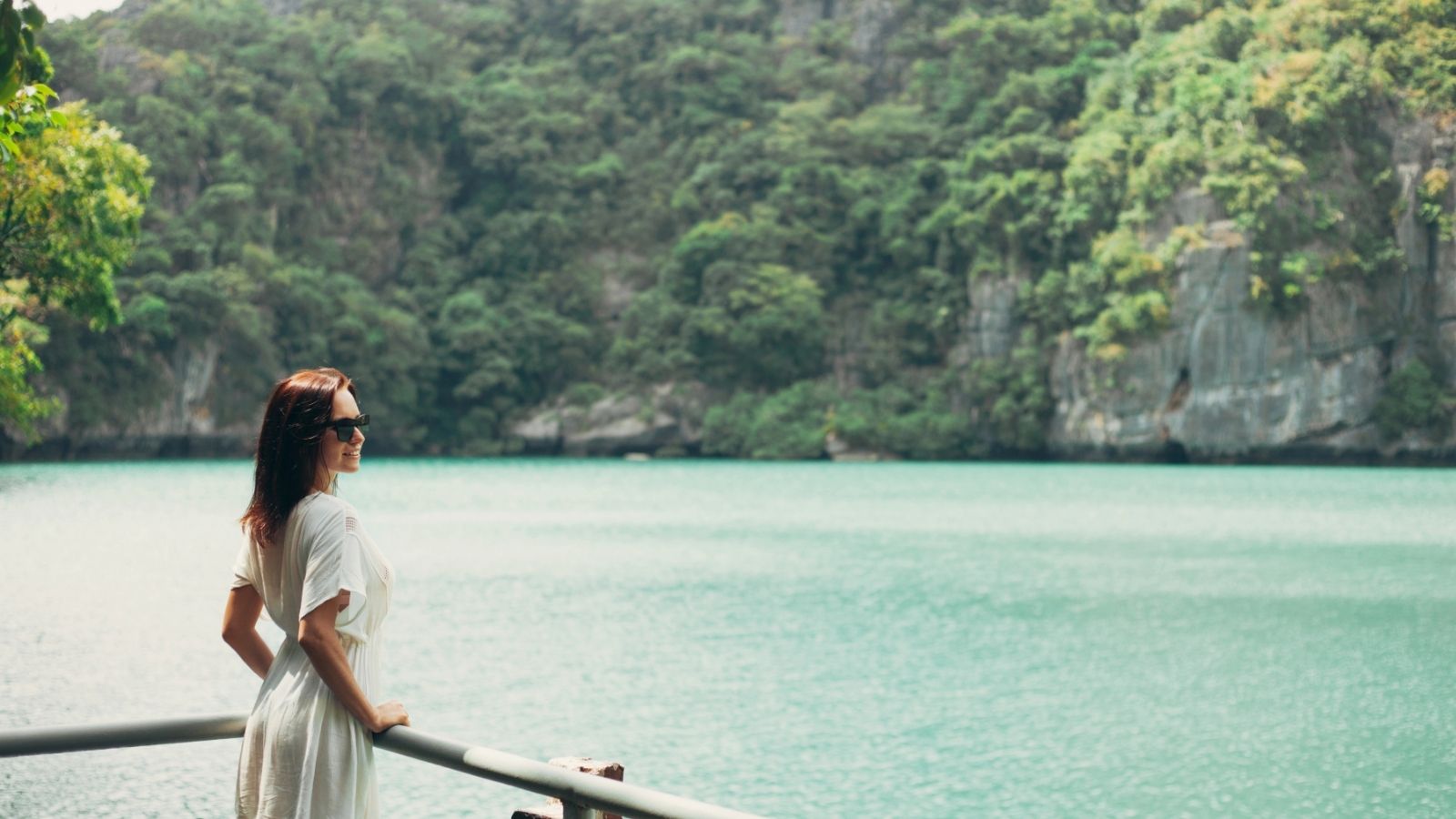 Woman in white dress and sunglasses standing by a railing, overlooking a turquoise lake surrounded by green trees—a perfect moment for hot weather travel on your next Asia trip.