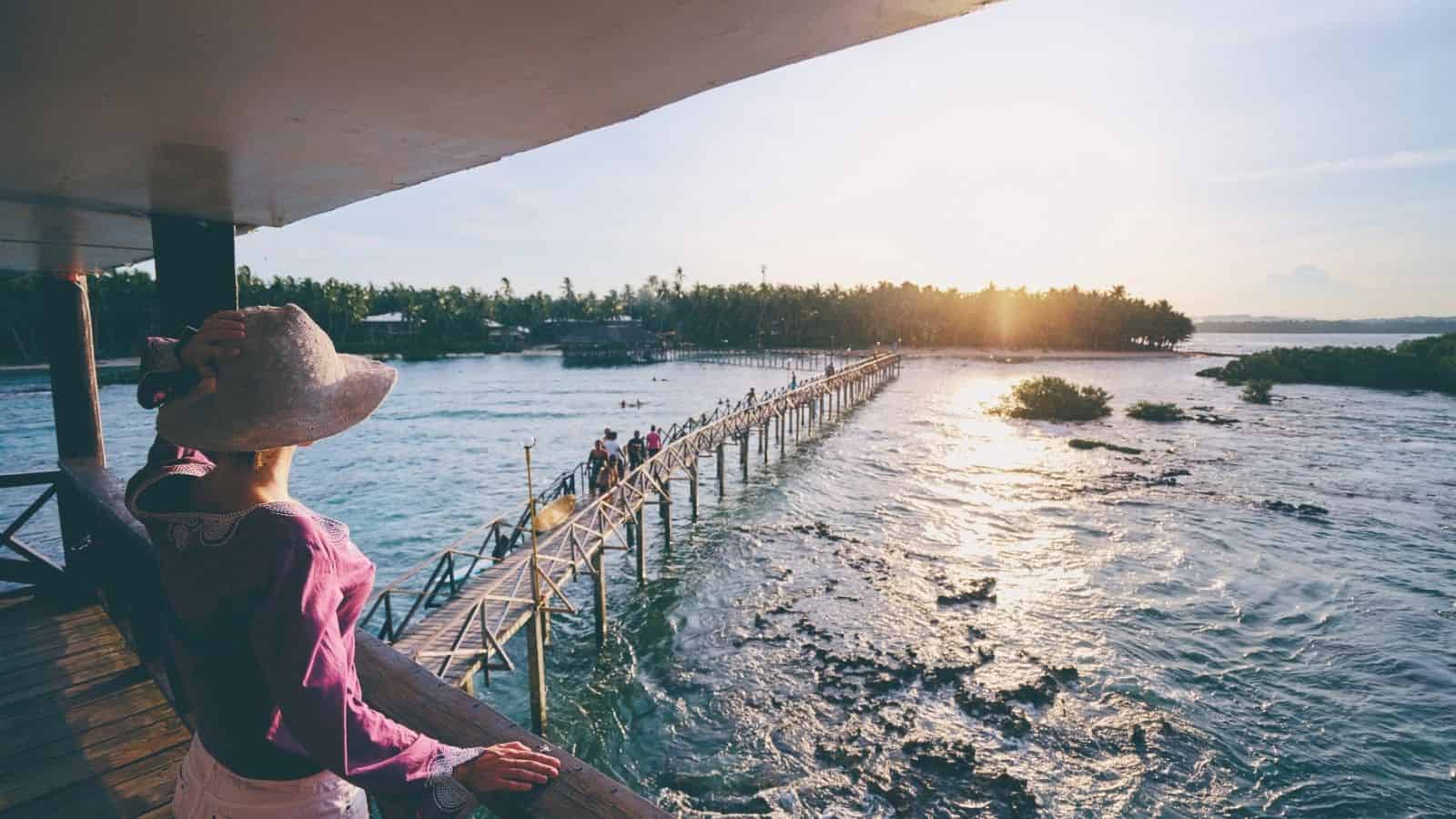 Person in hat looks out over a wooden bridge stretching across water toward trees at sunset.