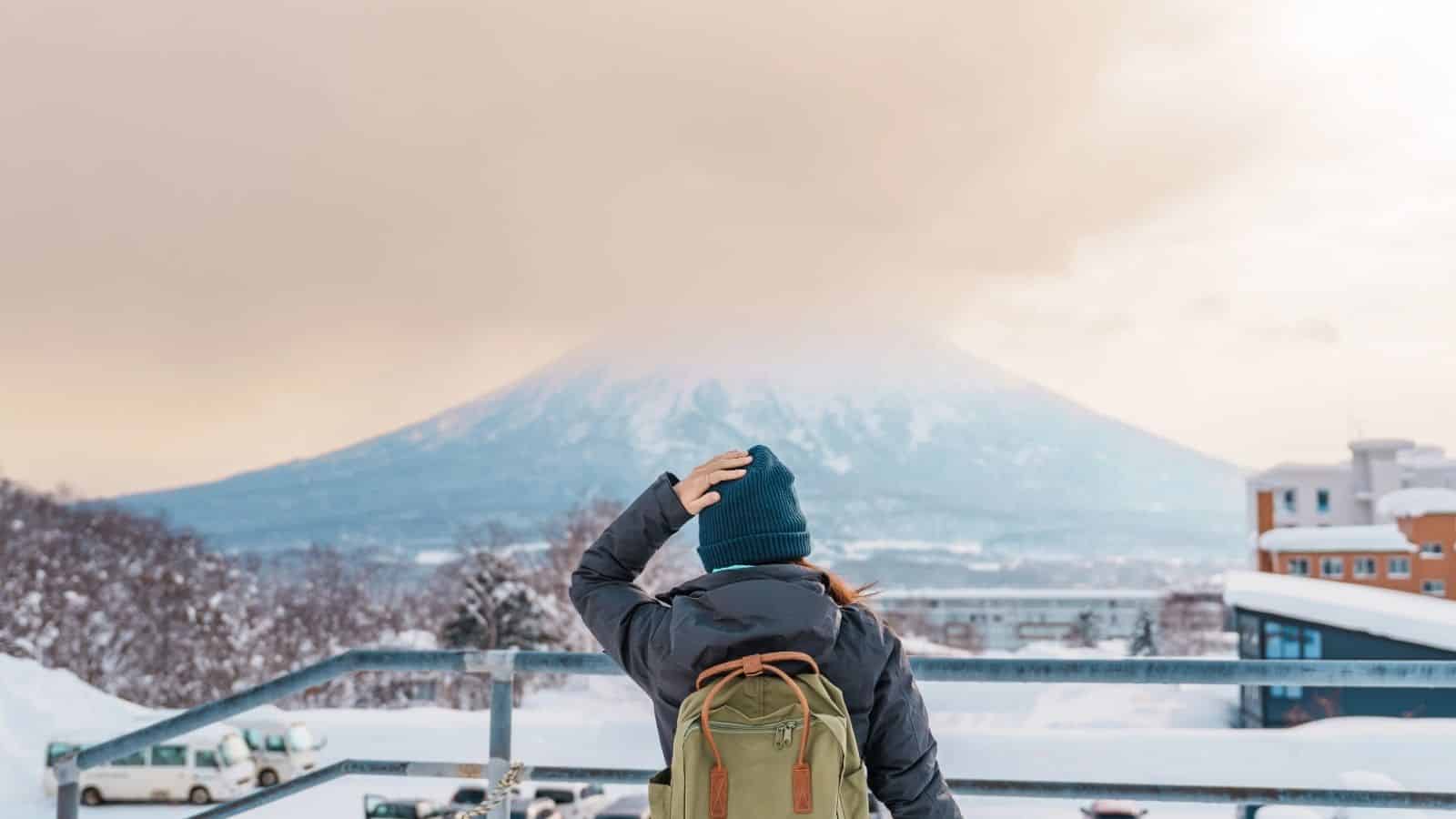 Person with a backpack looks at a snow-covered mountain, with cloudy skies and buildings in the foreground.