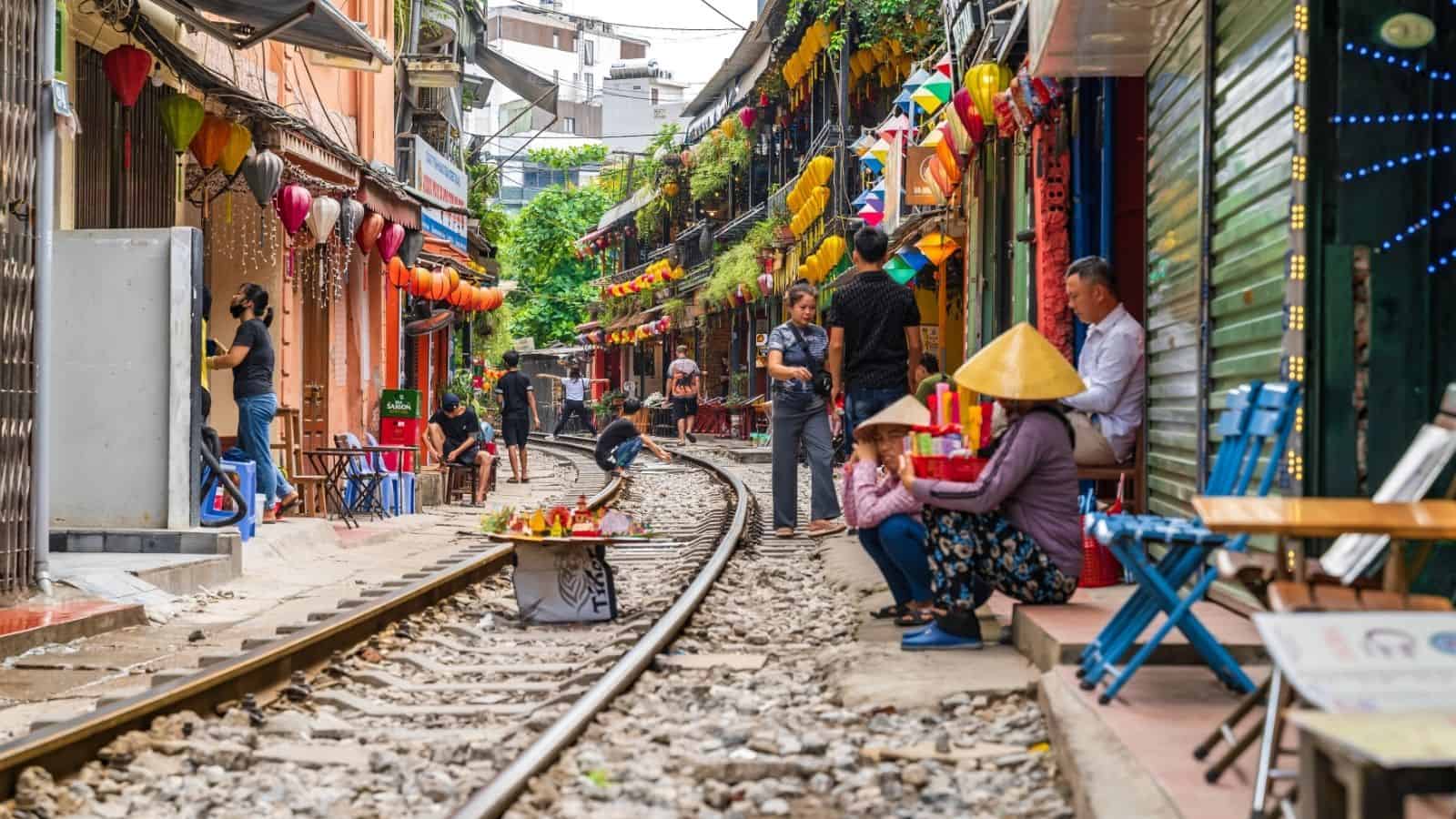 Narrow street with railway tracks, colorful lanterns, and people sitting or walking alongside shops and cafes.