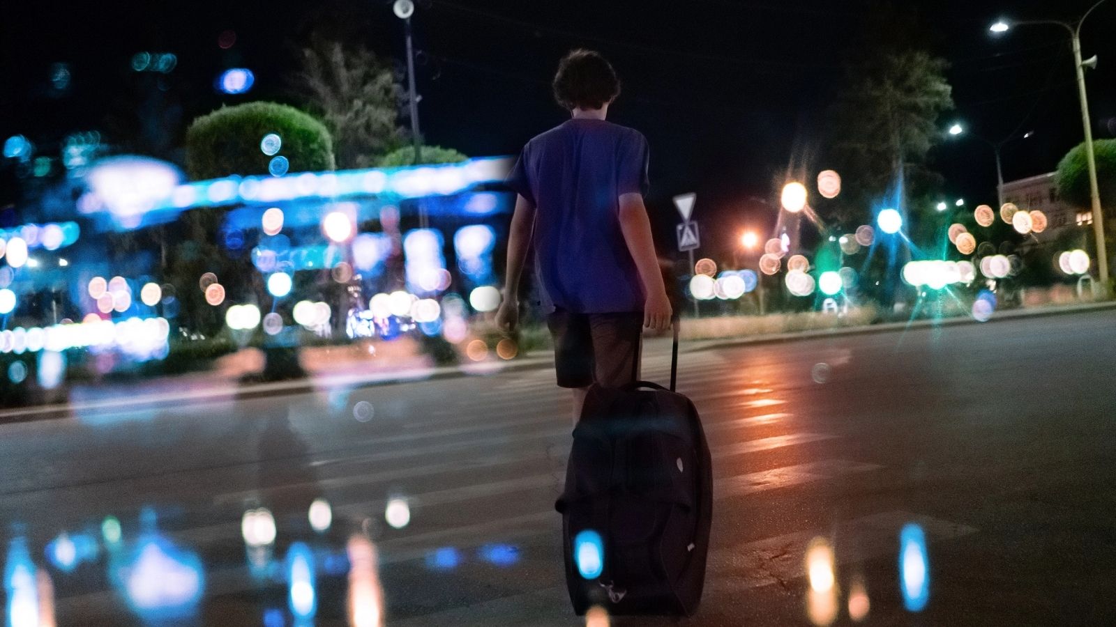 Person pulling a suitcase across a city street at night, with colorful bokeh lights in the background.