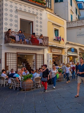 Bustling European street with people walking, shopping, and dining at outdoor cafes on a sunny day.