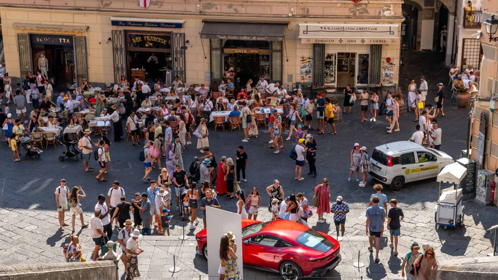 Busy Italian square with crowds, cafes, a parked red sports car, and a white van on a sunny day.