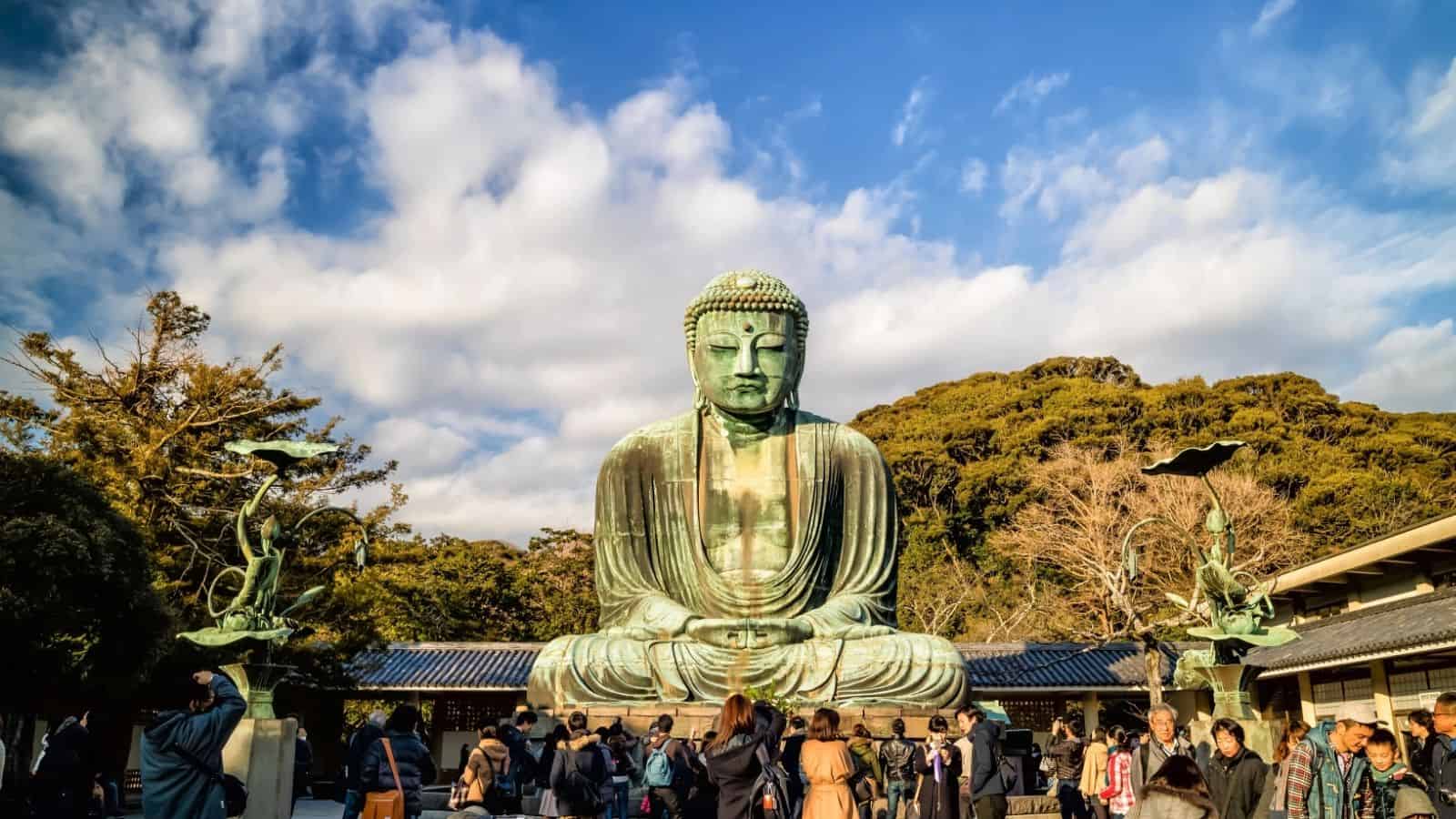 A large outdoor Buddha statue with people gathered in front, trees, and a blue sky with clouds in the background makes this one of the serene Japan summer destinations.