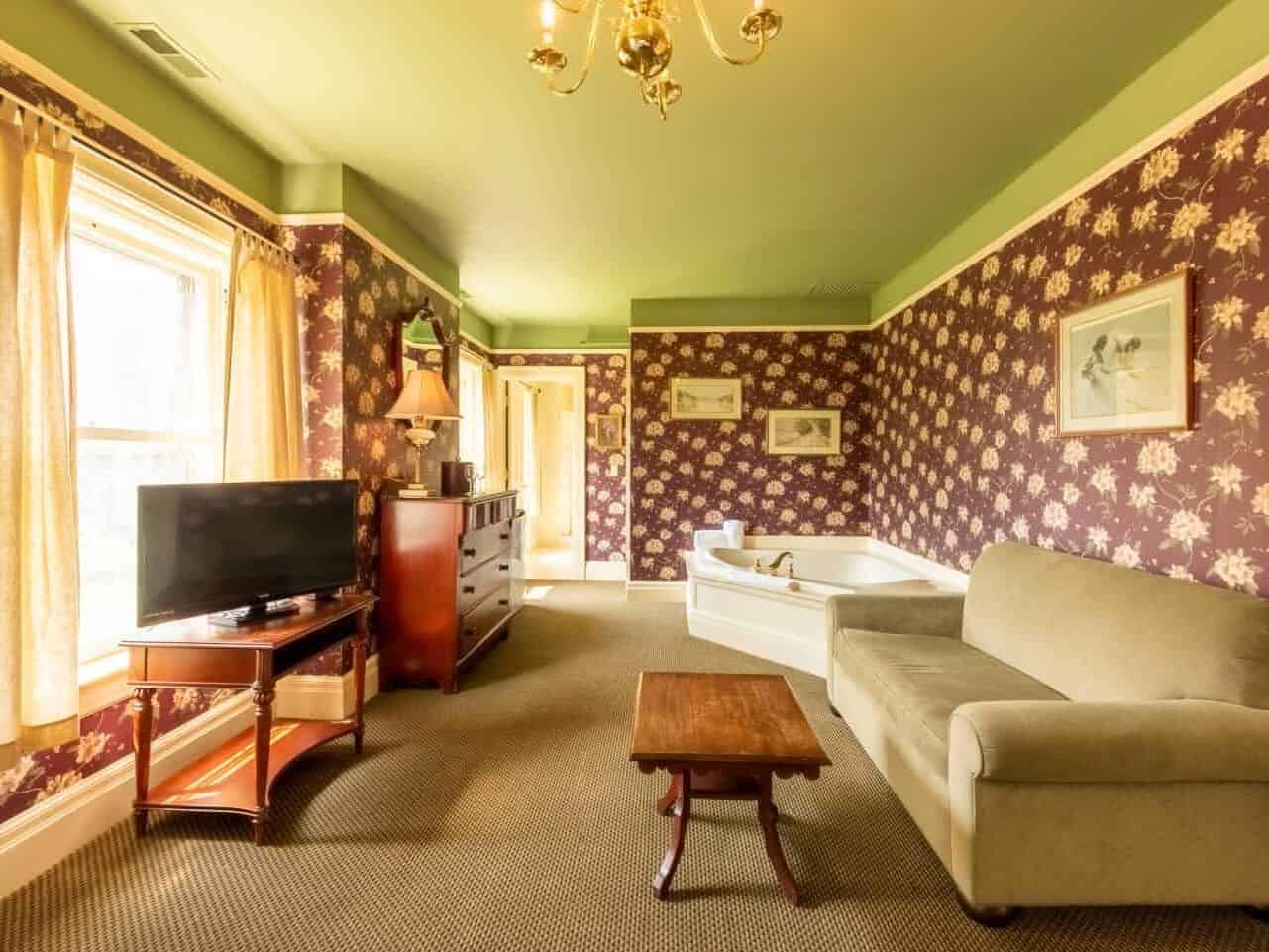 A cozy room with floral wallpaper, a green sofa, TV, dresser, and bathtub by a window with yellow curtains&mdash;perfect for relaxing after exploring nearby Finger Lakes wineries with lodging.
